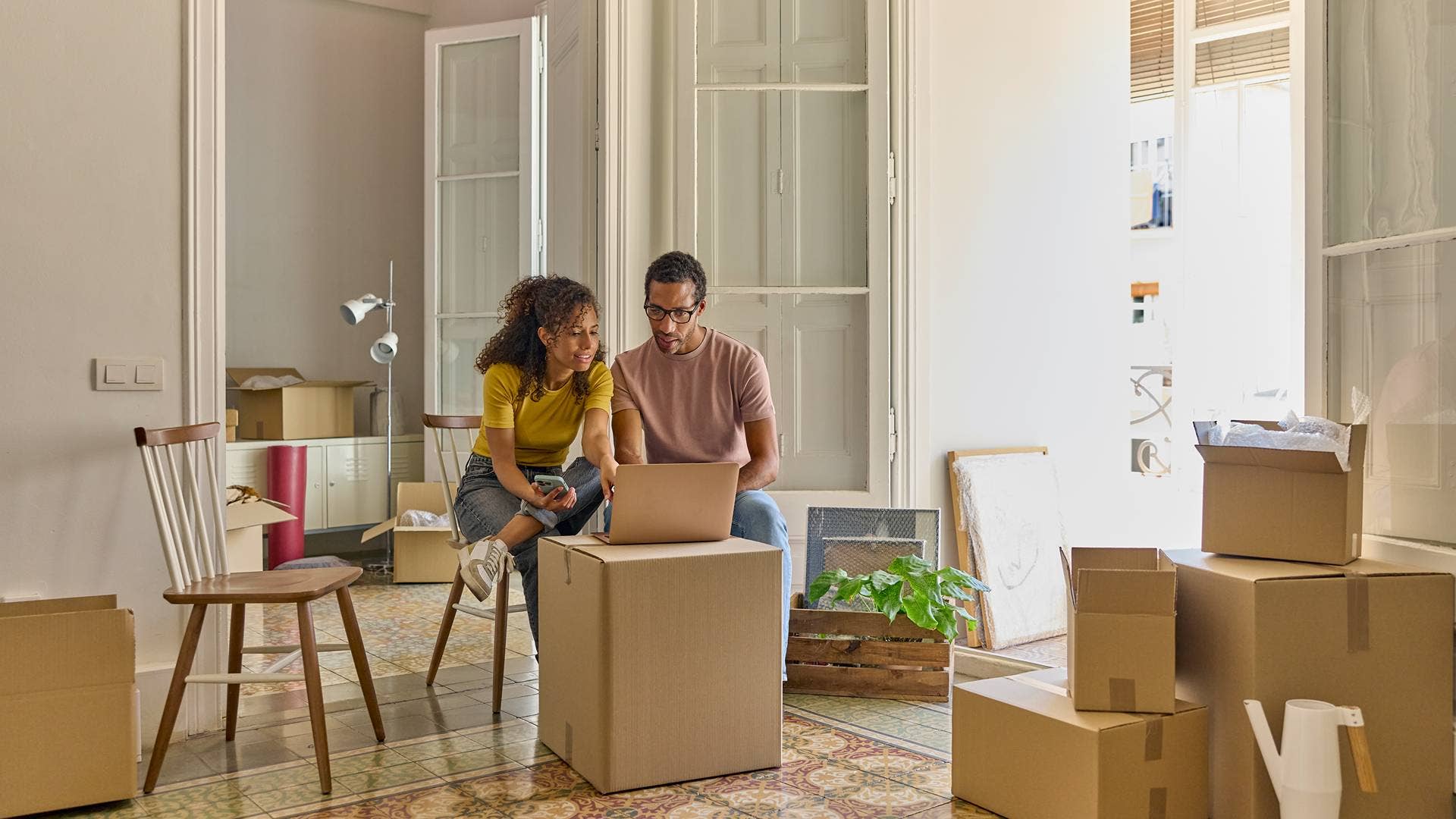 A young couple sit among moving boxes, looking at a laptop computer.