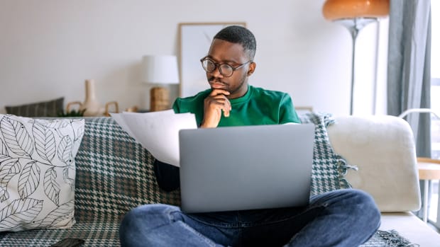Young casually clothed concentrated man going over paperwork while working from living room, he is sitting on the sofa with laptop in his lap