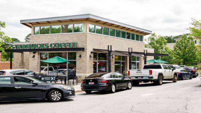 A Starbucks building exterior with a line of cars at the drive thru. Lead.