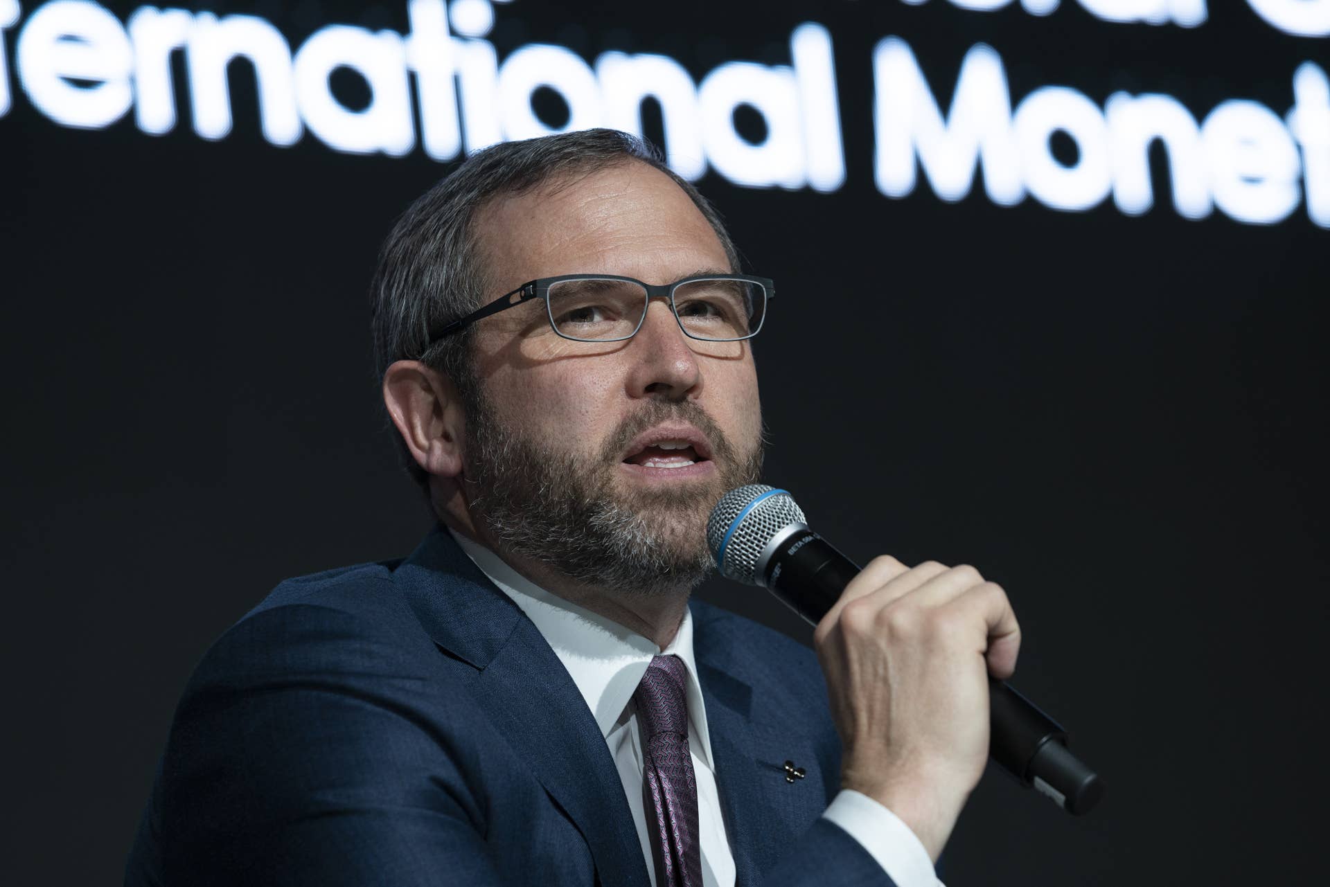 Brad is holding a microphone, wearing a grey suit and tie, speaking. He is wearing glasses.